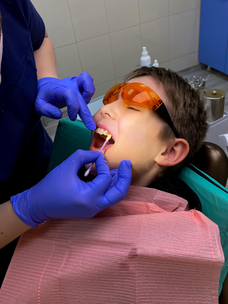 A young boy wearing orange protective glasses having a treatment applied to his teeth with a swab by a dentist.