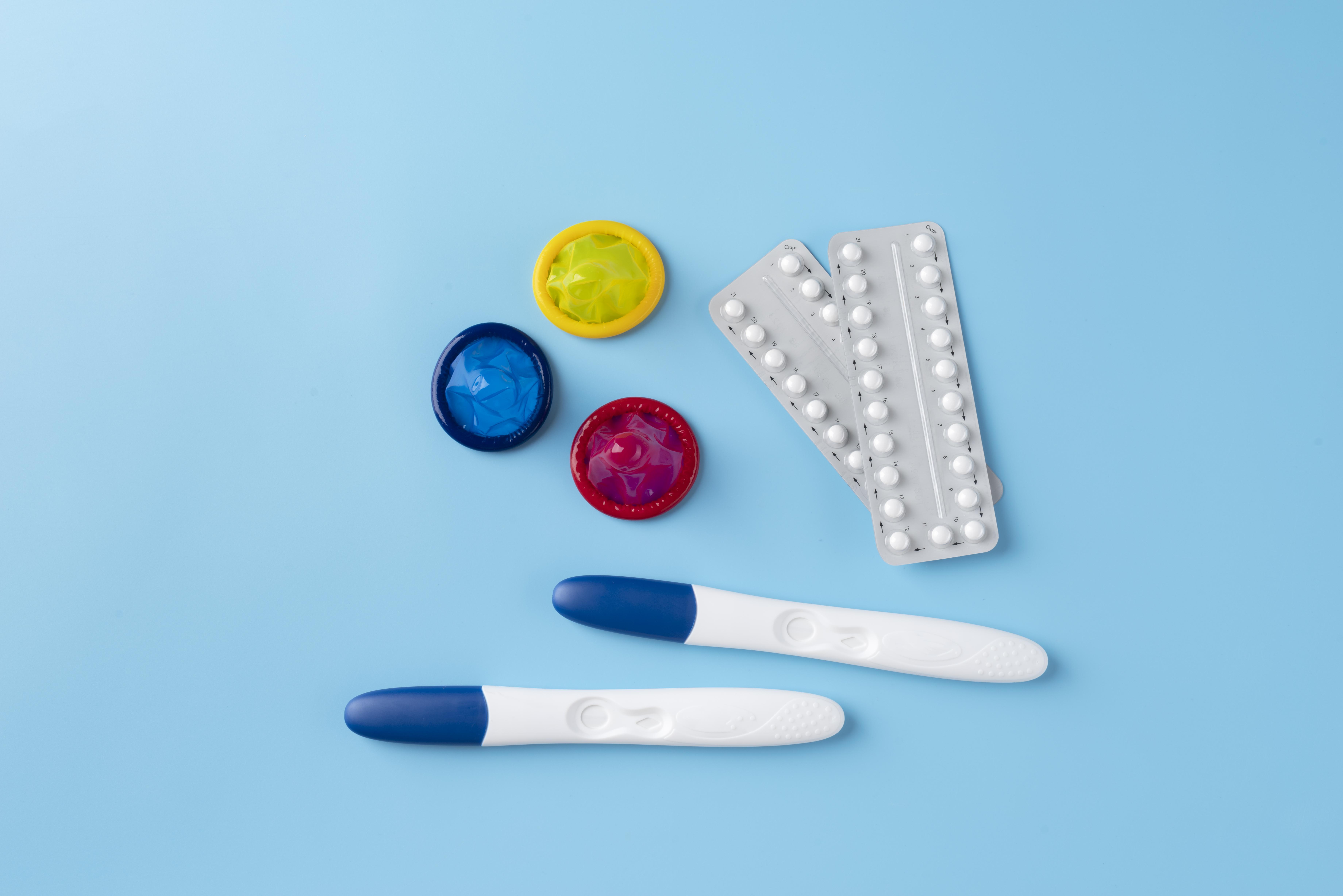 A flat lay arrangement of sexual health items including colorful condoms, birth control pills, and pregnancy tests on a blue background.